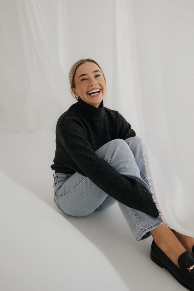 Lachende Frau im grauen Strickpullover sitzt auf dem Studioboden