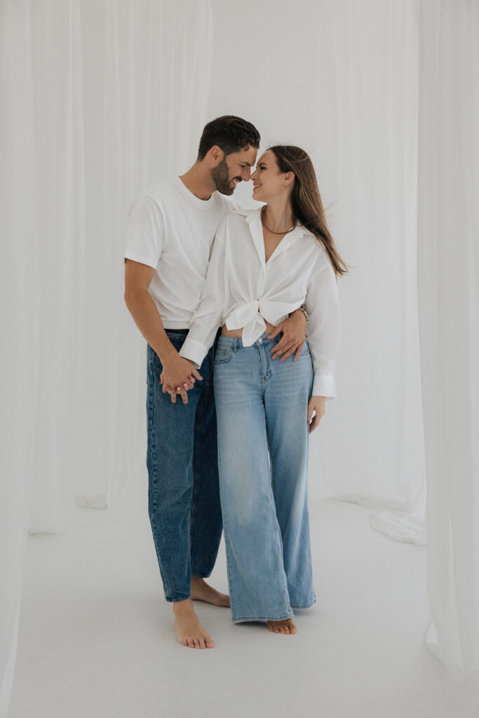 Paar in weißen Oberteilen und Jeans steht barfuß im hellen Studio